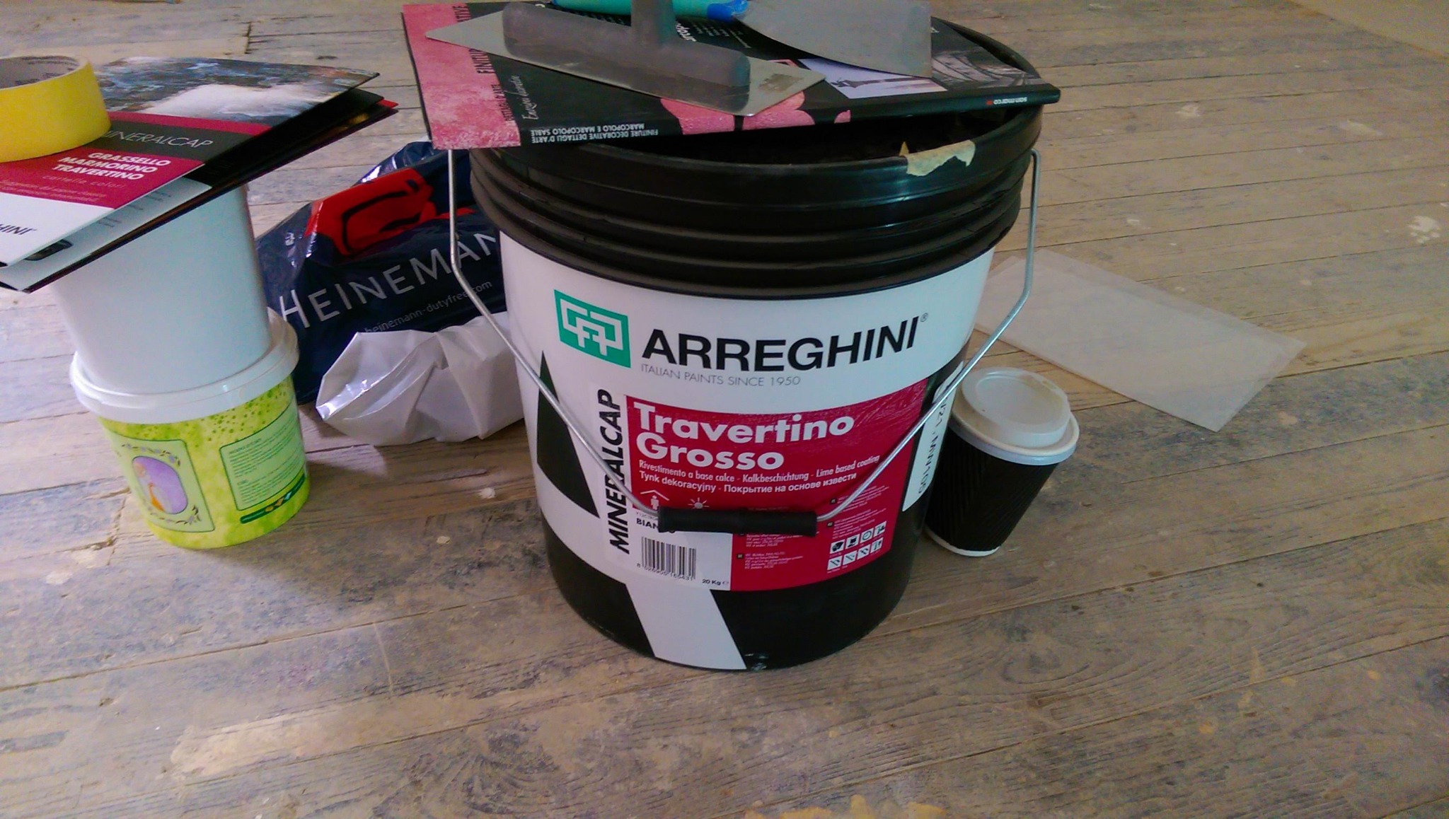 Wiadro z tynkiem dekoracyjnym Arreghini Travertino Grosso stoi na podłodze z paneli, obok kubek z kawą, puszka z żółtą taśmą malarską, katalogi i szpachelki.
