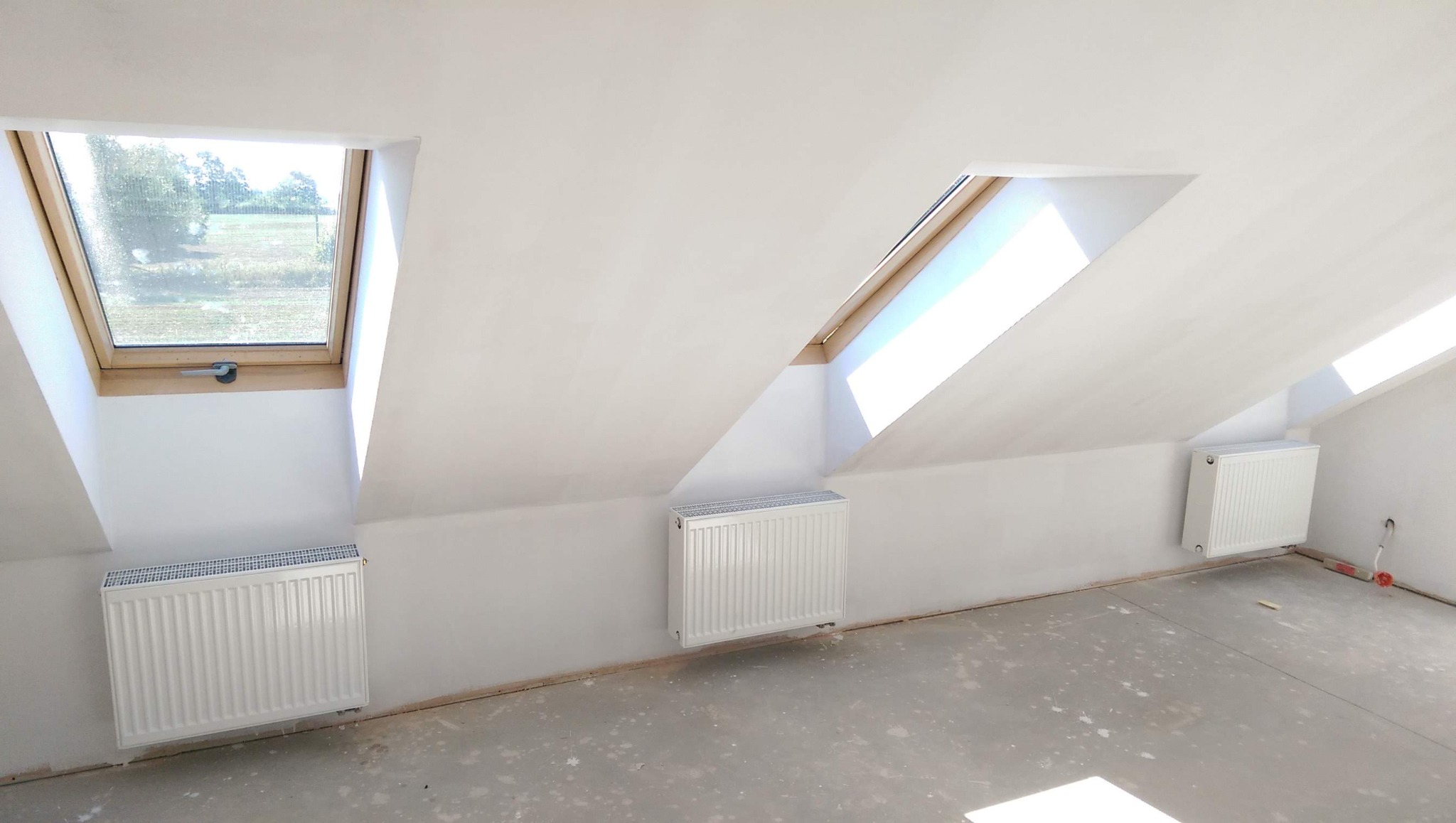 Poddasze ze świeżo pomalowanymi na biało ścianami, trzema oknami dachowymi i grzejnikami, podłoga przygotowana pod dalsze prace wykończeniowe.