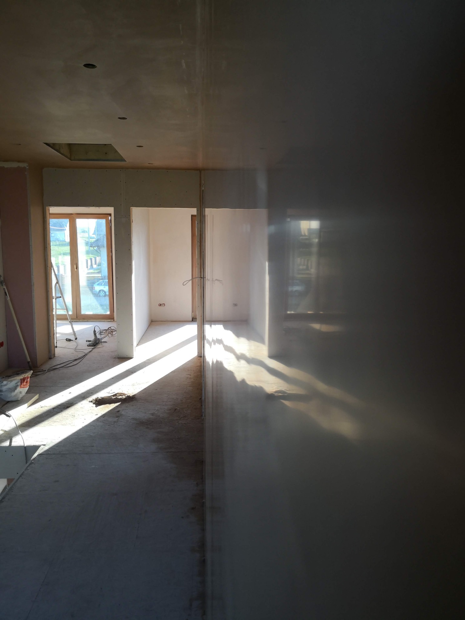 Wnętrze w trakcie prac wykończeniowych: surowe ściany, widoczne płyty kartonowo-gipsowe, szpachlowane powierzchnie, wpadające promienie słoneczne przez okno, drabina oparta o ścianę.