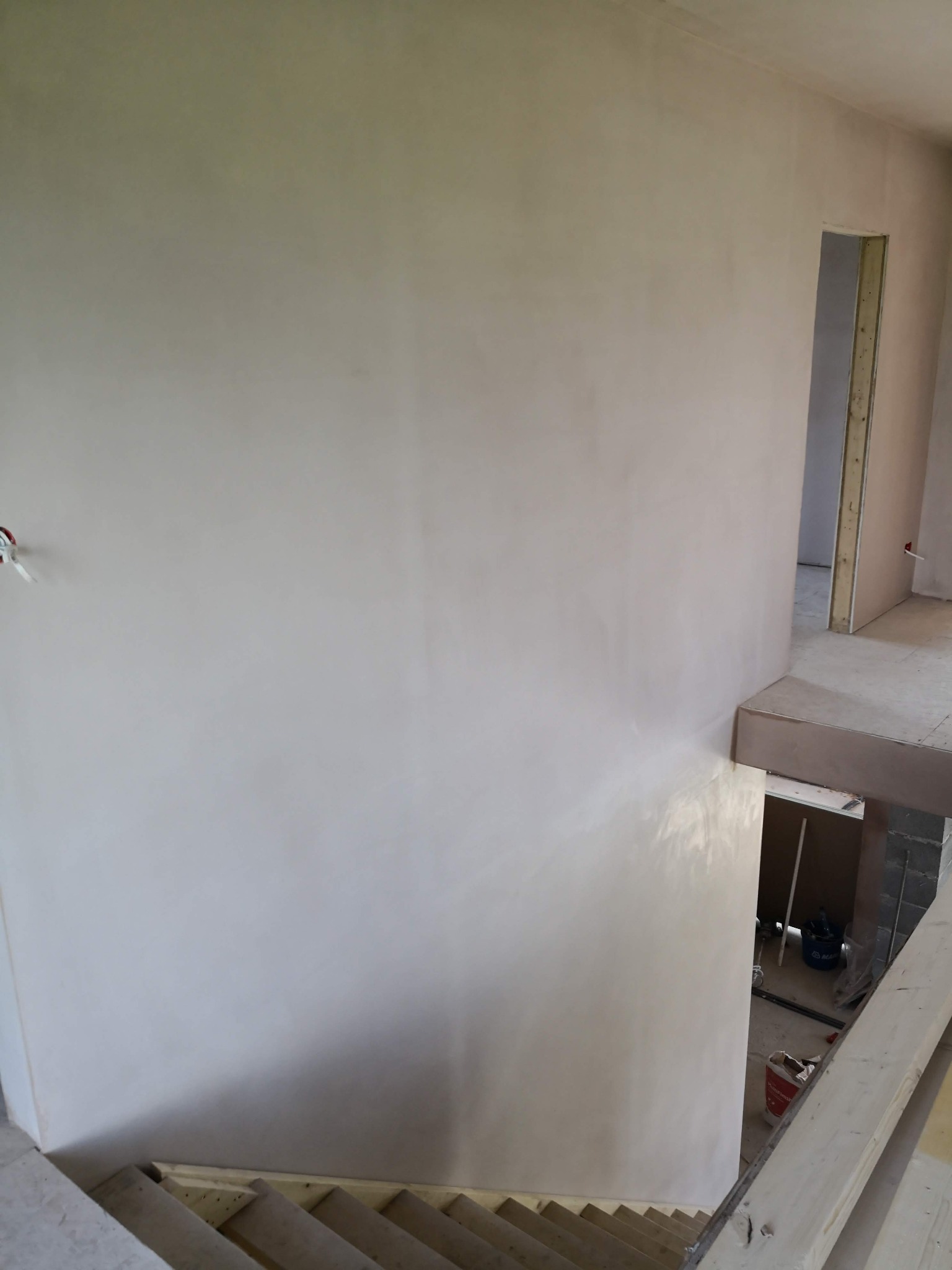 Świeżo wygładzona ściana w jasnym odcieniu w nowym domu we Wrocławiu, widok z góry na schody z surowego drewna i otwarte przejście do innego pomieszczenia w tle.