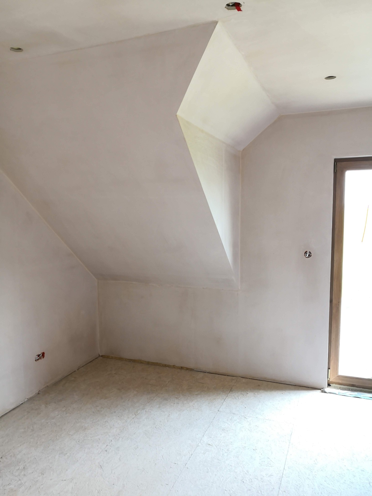 Pokój na poddaszu po szpachlowaniu, ściany i skośny sufit pokryte jasną masą, widoczne gniazdka elektryczne i otwór na oświetlenie sufitowe.