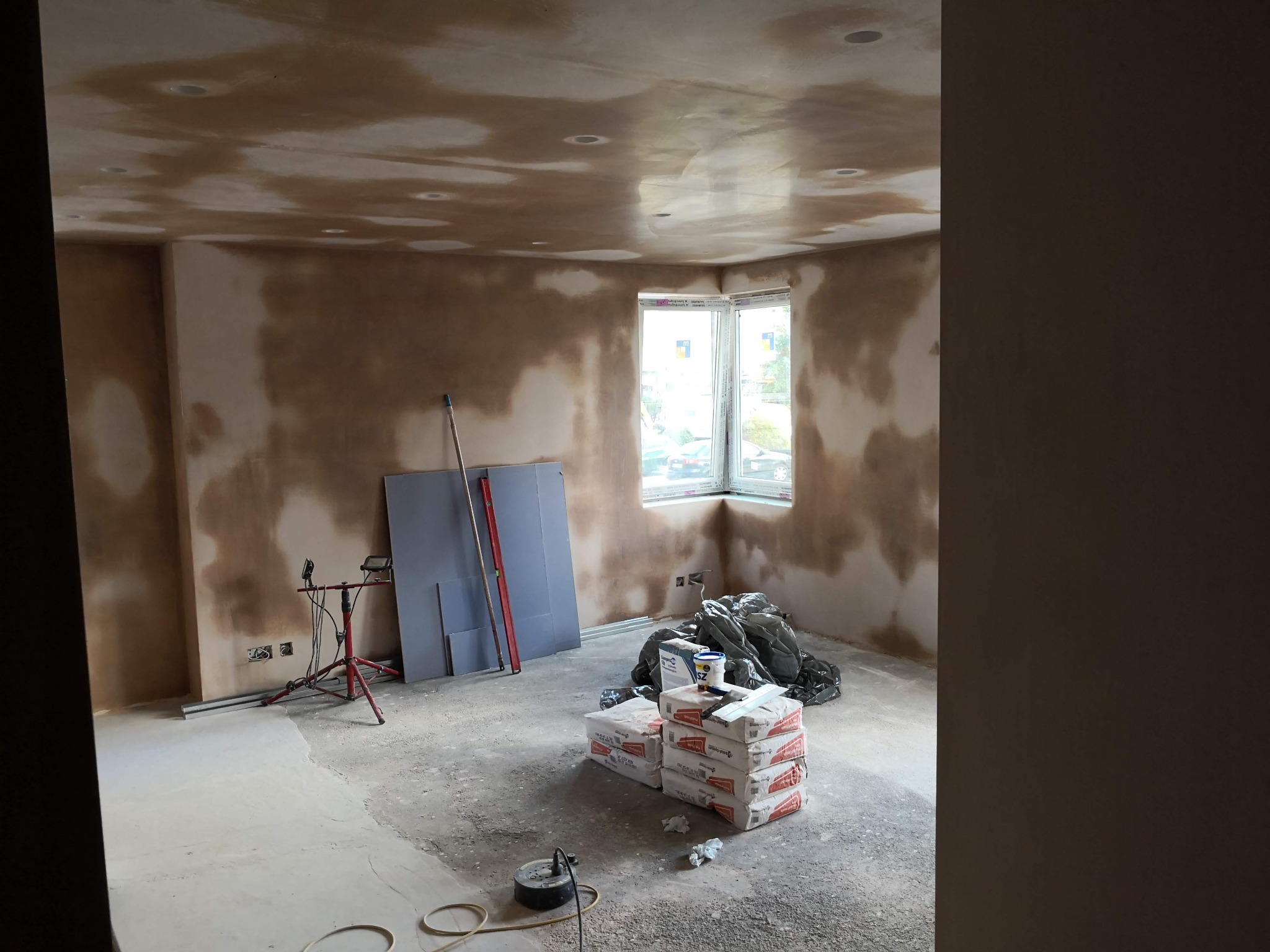 Pokój w trakcie prac wykończeniowych: ściany i sufit pokryte wstępną warstwą szpachli, widoczne worki z materiałami, poziomica oparta o ścianę, lampa na statywie i okno z widokiem na zewnątrz.