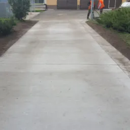 Świeżo wylana, betonowa droga dojazdowa z widocznym odpływem liniowym i pracownikami w odblaskowych kamizelkach w tle.