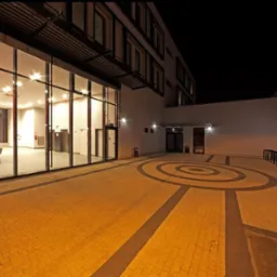 Oświetlony nocą dziedziniec z kostki brukowej z geometrycznym wzorem, przylegający do nowoczesnego budynku z przeszkloną fasadą i metalową konstrukcją zadaszenia.