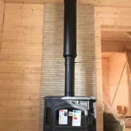 Czarny piec wolnostojący z długim kominem, zainstalowany przy ścianie z cegły i boazerii w surowym wnętrzu z widoczną konstrukcją dachu.