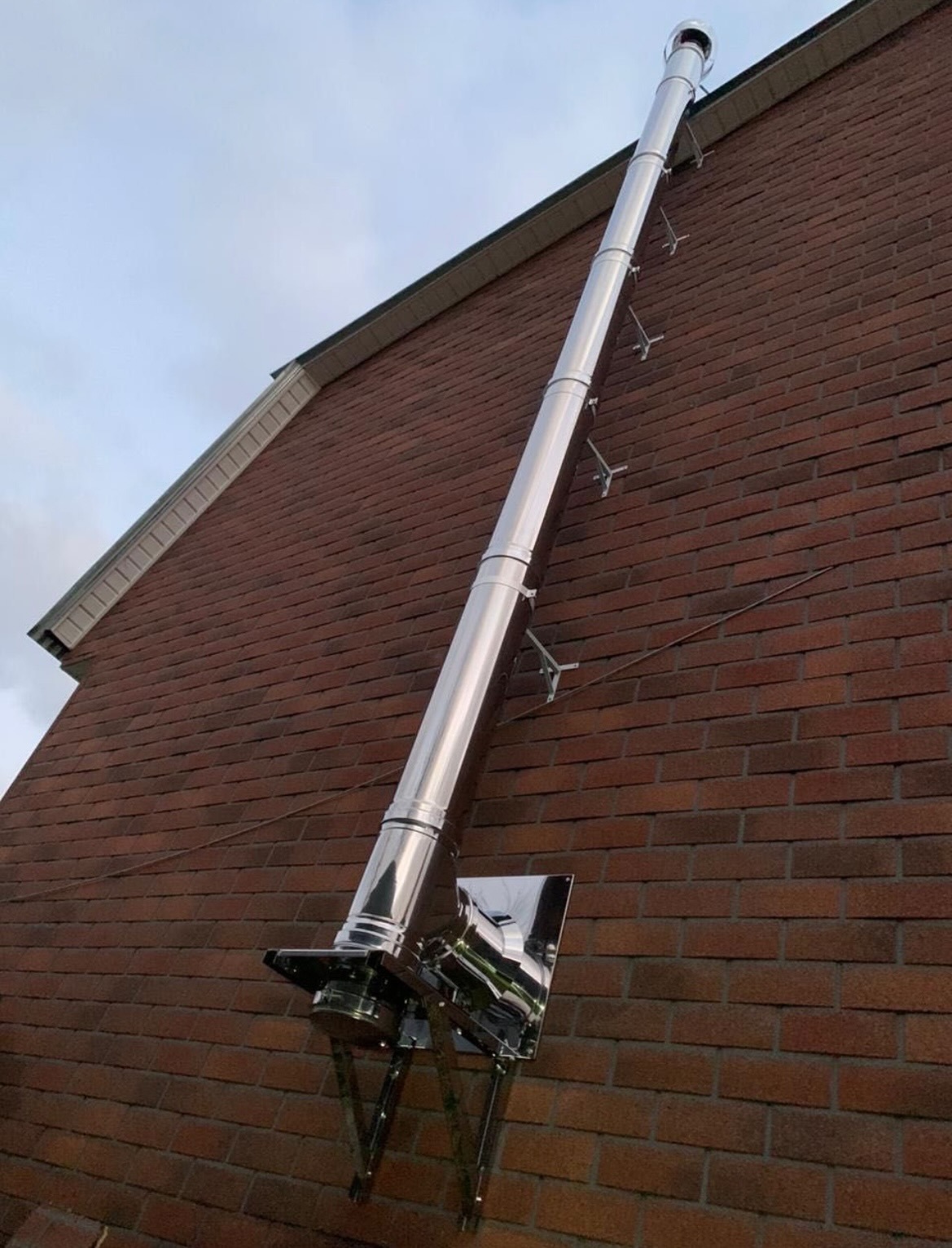 Zewnętrzny, chromowany komin zamontowany na ceglanej ścianie budynku z widocznymi uchwytami mocującymi i metalową podstawą.