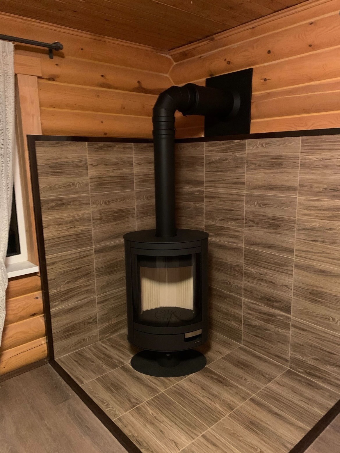 Czarny piec wolnostojący z rurą odprowadzającą spaliny, zamontowany w rogu pomieszczenia wyłożonego płytkami imitującymi drewno, Białystok.