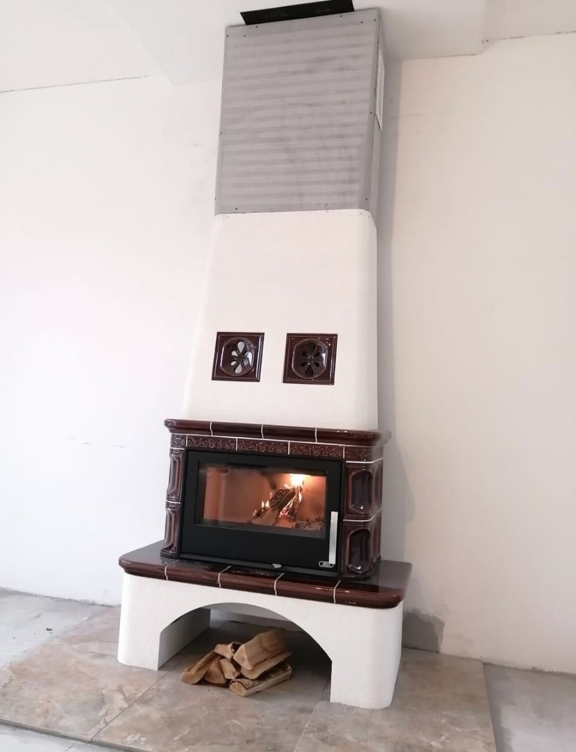 Kaflowy kominek z palącym się drewnem, widoczne drewno pod paleniskiem, białe ściany, jasna podłoga.