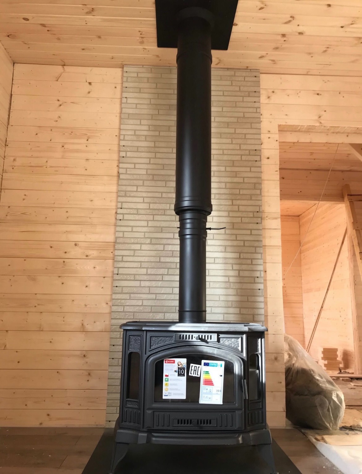 Czarny piec wolnostojący z długim kominem, zainstalowany przy ścianie z cegły i boazerii w surowym wnętrzu z widoczną konstrukcją dachu.
