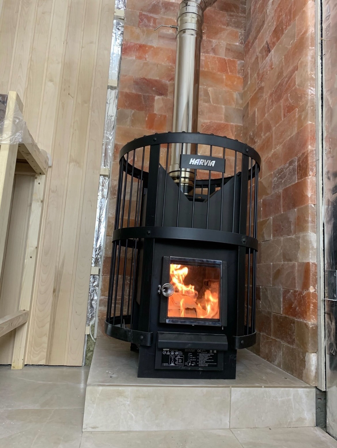 Czarny piec Harvia z płomieniami widocznymi przez szybę, obudowany metalową klatką, z kominem ze stali nierdzewnej, na tle ściany z różowej cegły solnej i drewnianej boazerii.