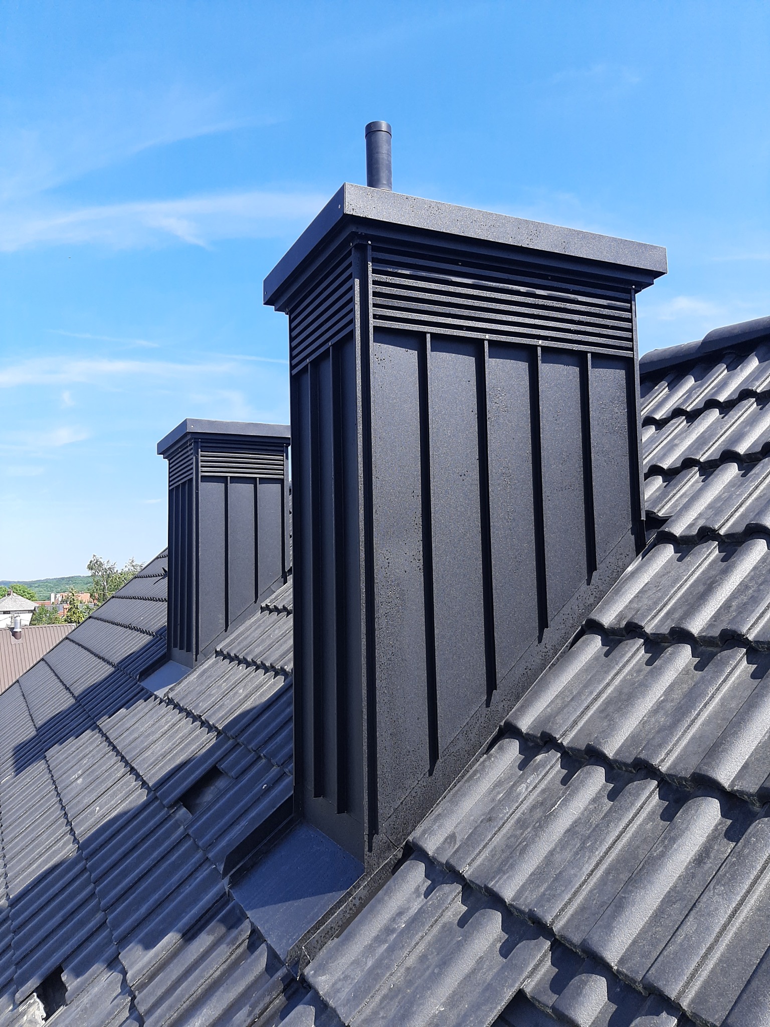 Dwa czarne kominy wentylacyjne na dachu z ciemnoszarej dachówki ceramicznej, błękitne niebo w tle.
