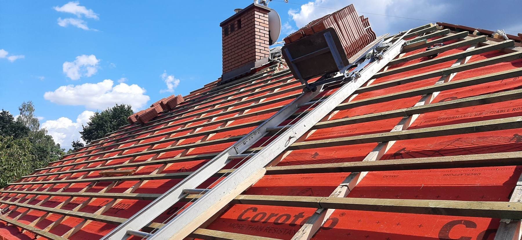 Konstrukcja dachu z łatami, membraną w kolorze czerwonym z widocznym kominem i podnośnikiem do transportu dachówek na tle błękitnego nieba.