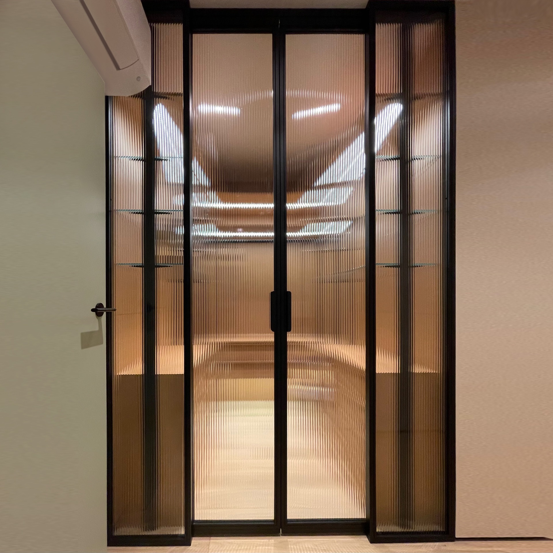 Dwuskrzydłowe drzwi przesuwne z ryflowanego szkła w czarnej ramie, widok na oświetlone wnętrze z półkami.