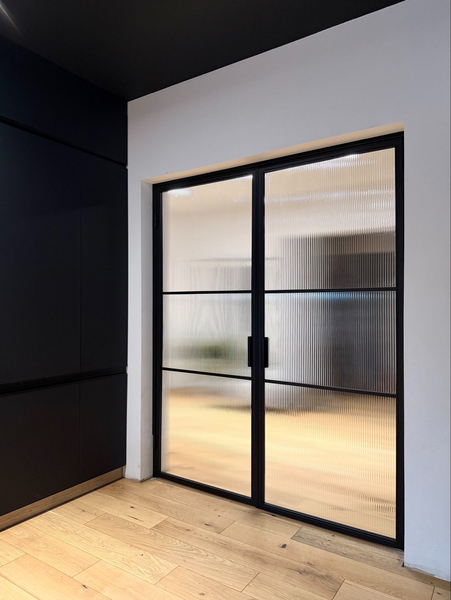 Nowoczesne dwuskrzydłowe drzwi z ryflowanego szkła w czarnej ramie, zamontowane w jasnym wnętrzu z drewnianą podłogą i czarnymi meblami.