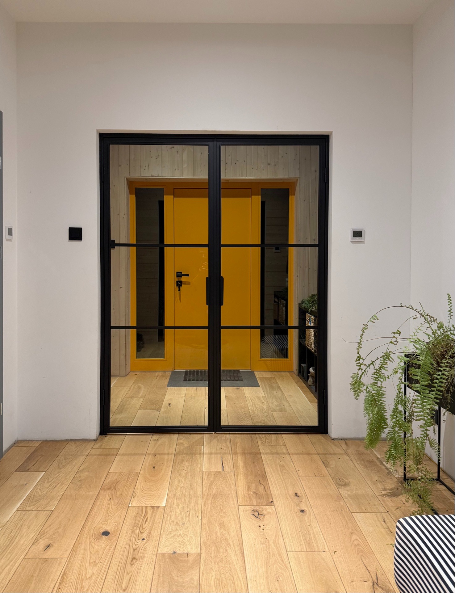 Dwuskrzydłowe drzwi z czarną ramą i podziałami szklanymi, widoczne na tle żółtych drzwi wejściowych i drewnianej podłogi