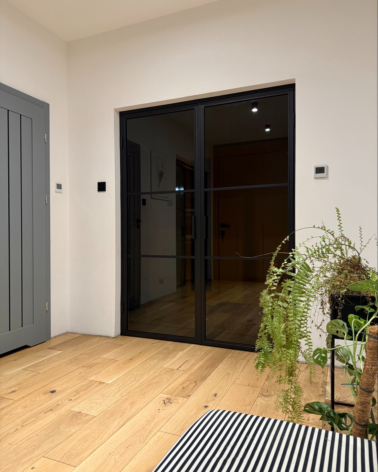 Nowoczesne, dwuskrzydłowe drzwi przesuwne z czarnymi szprosami i szklanym wypełnieniem, widoczne z perspektywy wnętrza, z drewnianą podłogą i roślinami doniczkowymi w tle.