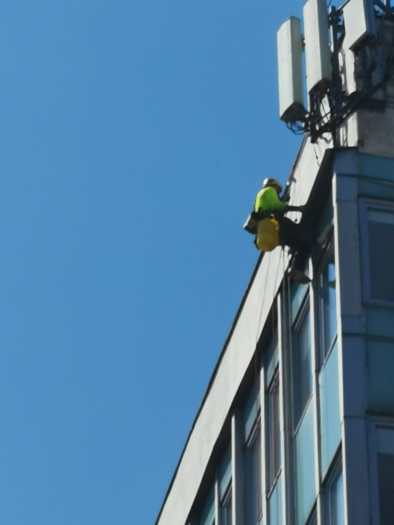 Pracownik w uprzęży, w jaskrawej kamizelce, na wysokości przy antenach telekomunikacyjnych na budynku biurowym z niebieską elewacją, widok z dołu.