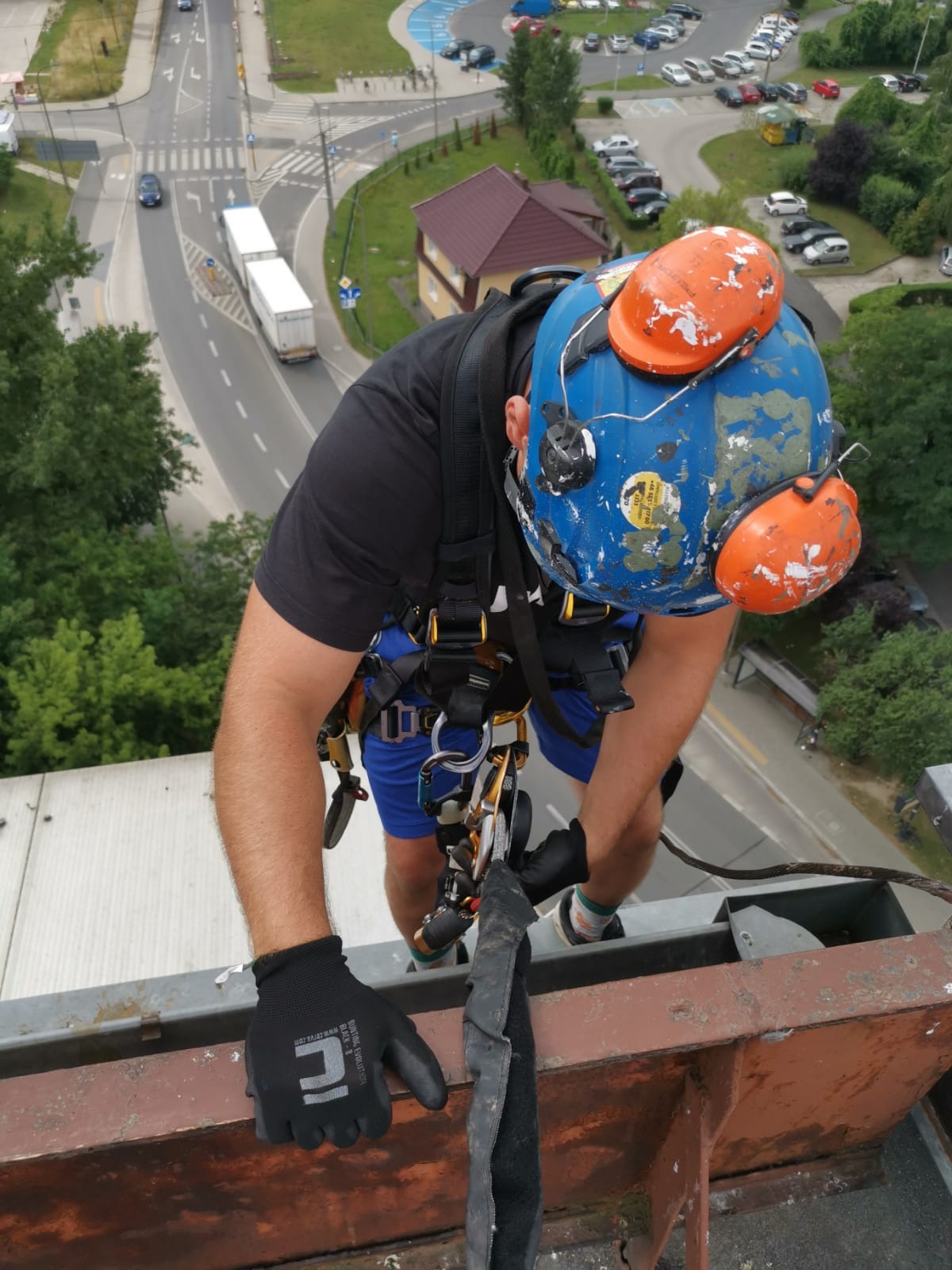 Pracownik wysokościowy w niebieskim hełmie z pomarańczowymi nausznikami, zabezpieczony uprzężą, wykonuje prace na wysokości, widok z góry na ulicę z ruchem samochodowym.