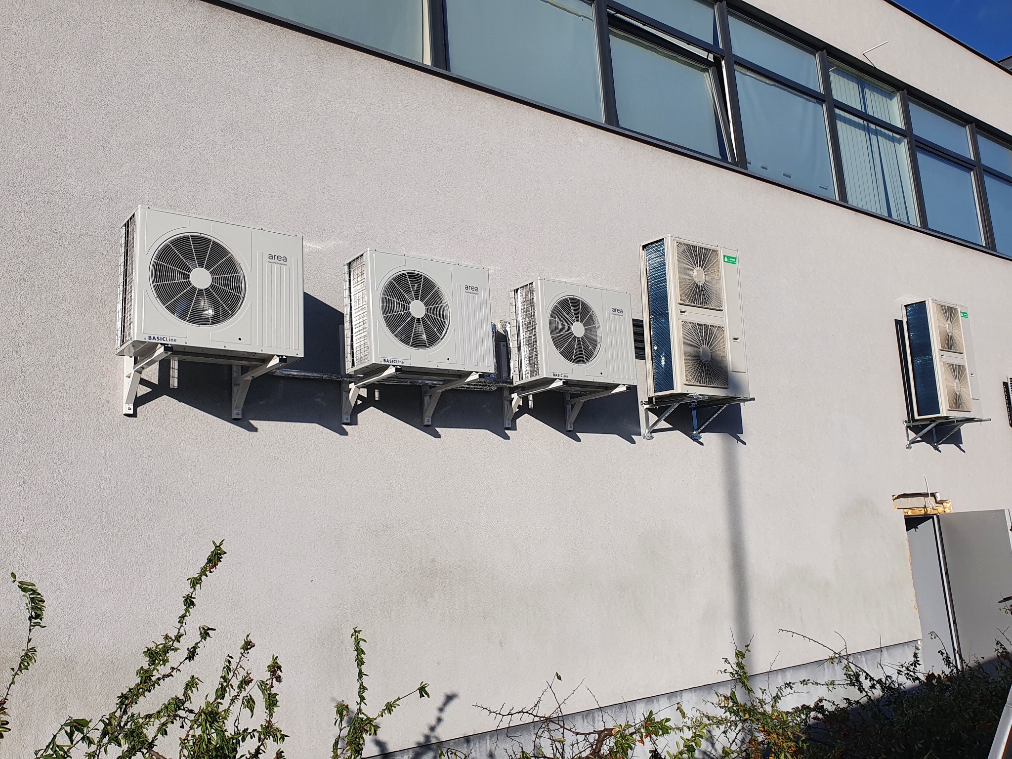Szereg zewnętrznych jednostek klimatyzacyjnych zamontowanych na jasnej ścianie budynku biurowego, widoczne okna i roślinność u podstawy ściany.