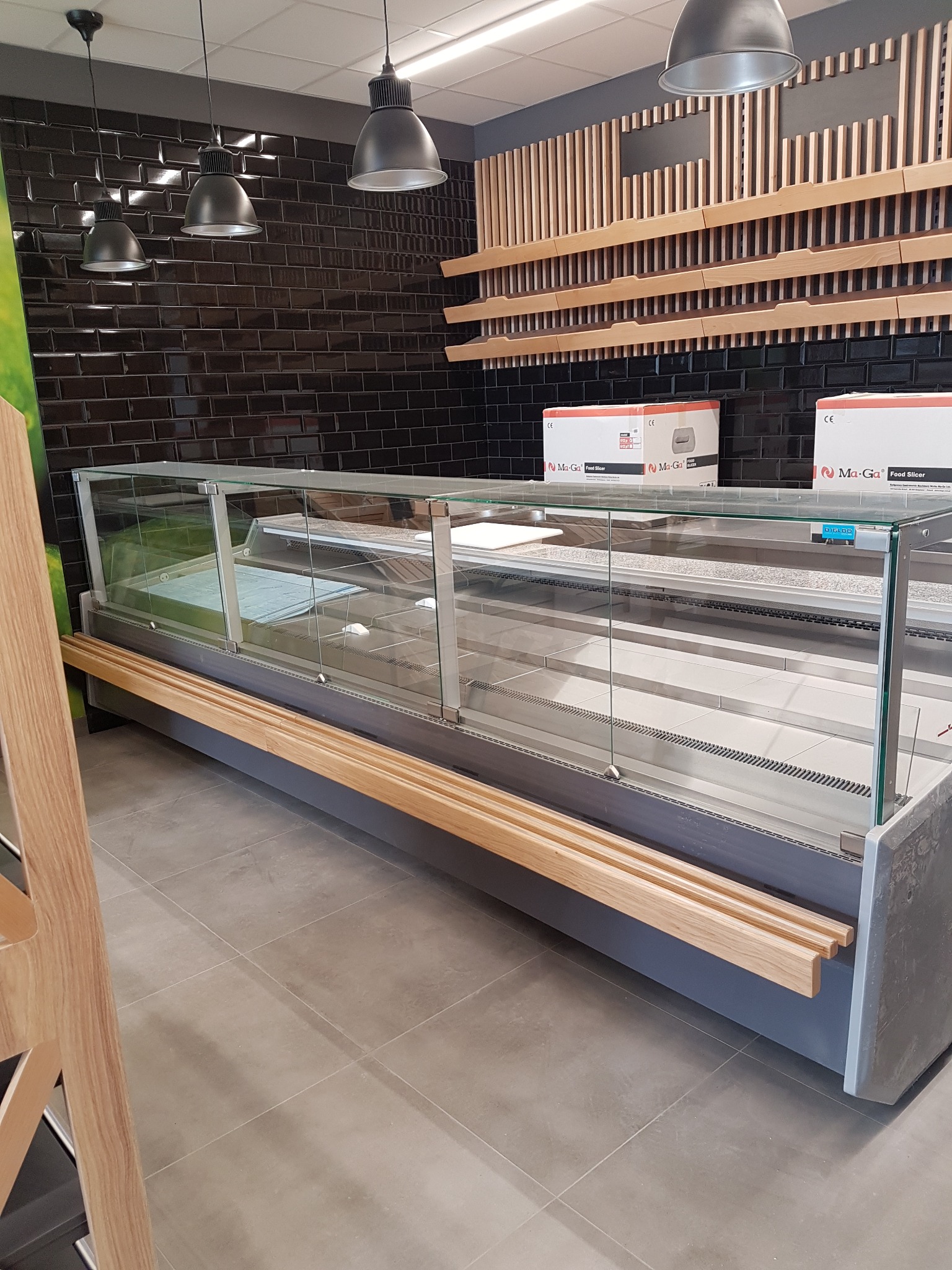 Pusta, przeszklona lada chłodnicza w sklepie z czarną, ceglaną ścianą w tle i drewnianymi elementami wystroju.