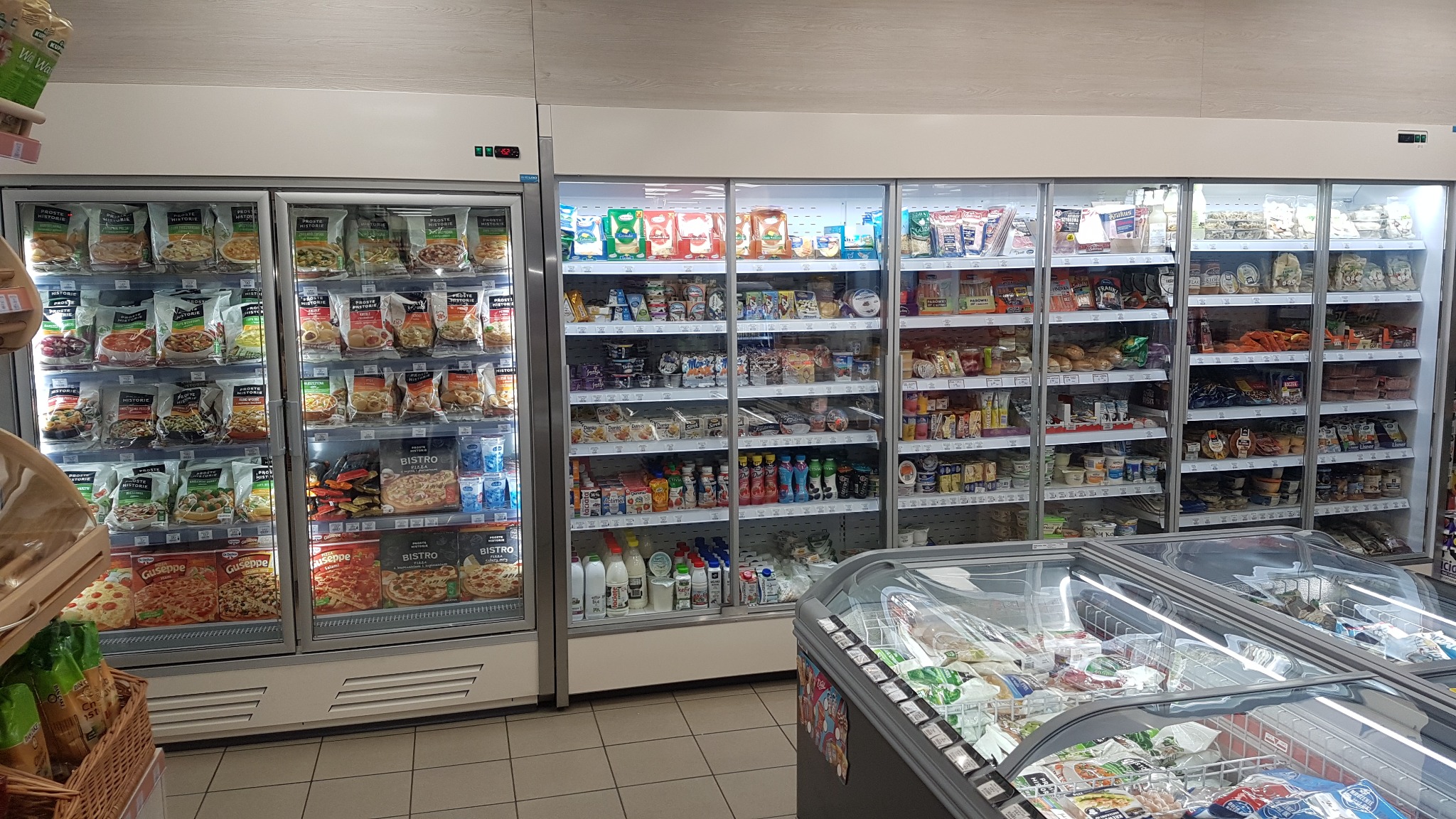Rząd chłodziarek sklepowych z przeszklonymi drzwiami, wypełnionych mrożonkami, nabiałem i innymi produktami spożywczymi, widoczne z perspektywy klienta.