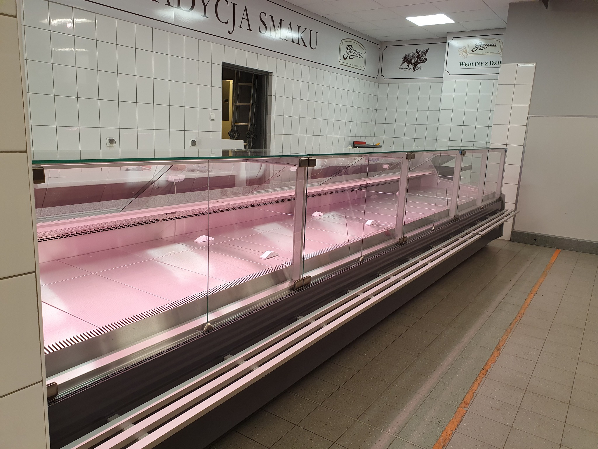 Pusta chłodnicza lada ekspozycyjna z oświetleniem LED, widok z perspektywy klienta, białe płytki na ścianach i logo producenta w tle.
