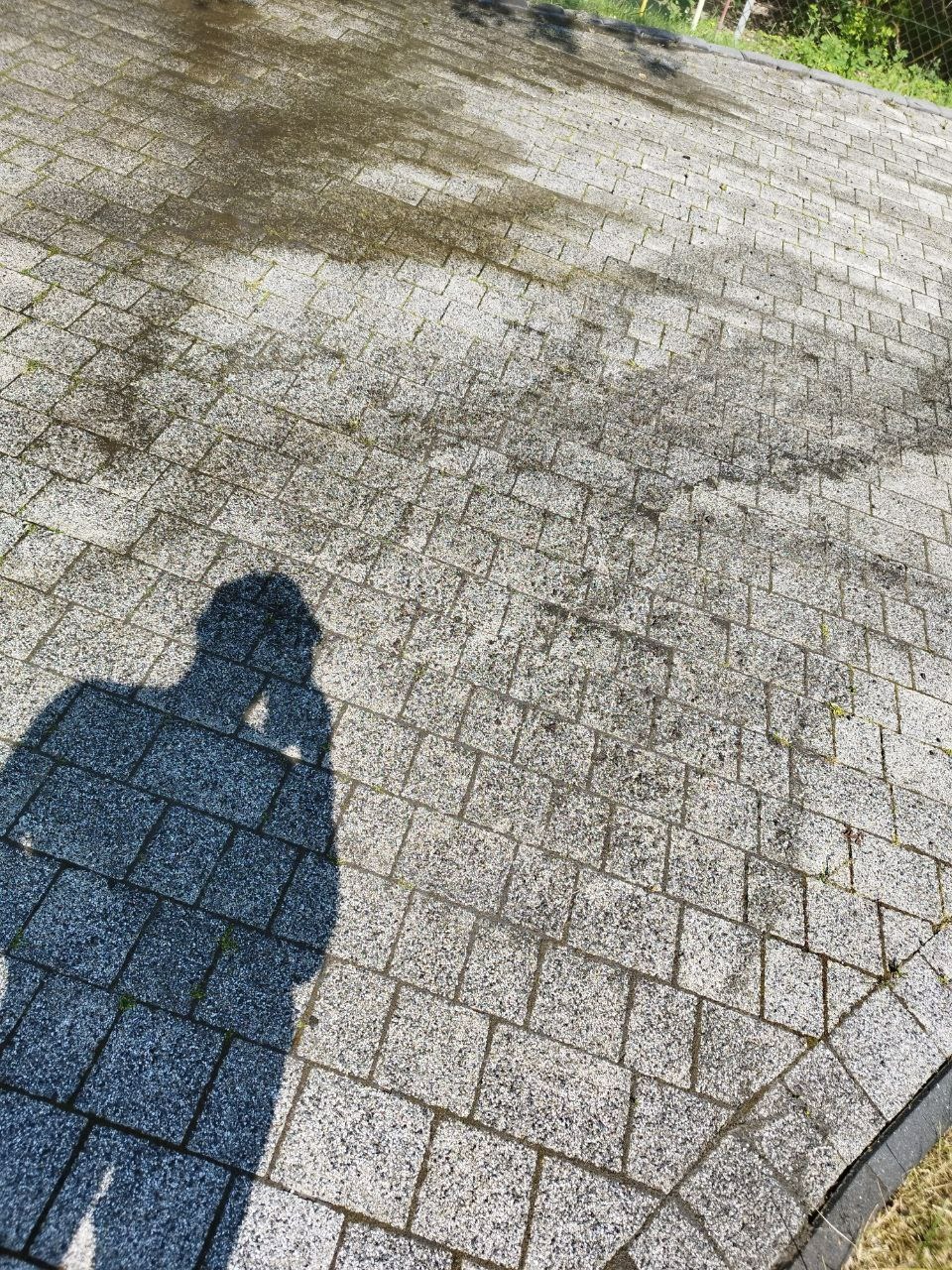 Częściowo umyta kostka brukowa z widocznym cieniem osoby fotografującej, ukazująca różnicę w czystości przed i po zabiegu.
