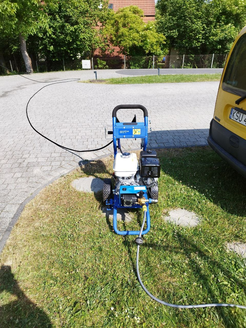 Profesjonalna myjka ciśnieniowa Nilfisk, gotowa do użycia w Katowicach, stoi na trawie obok chodnika, z podłączonym wężem. Widoczny fragment żółtego samochodu dostawczego.