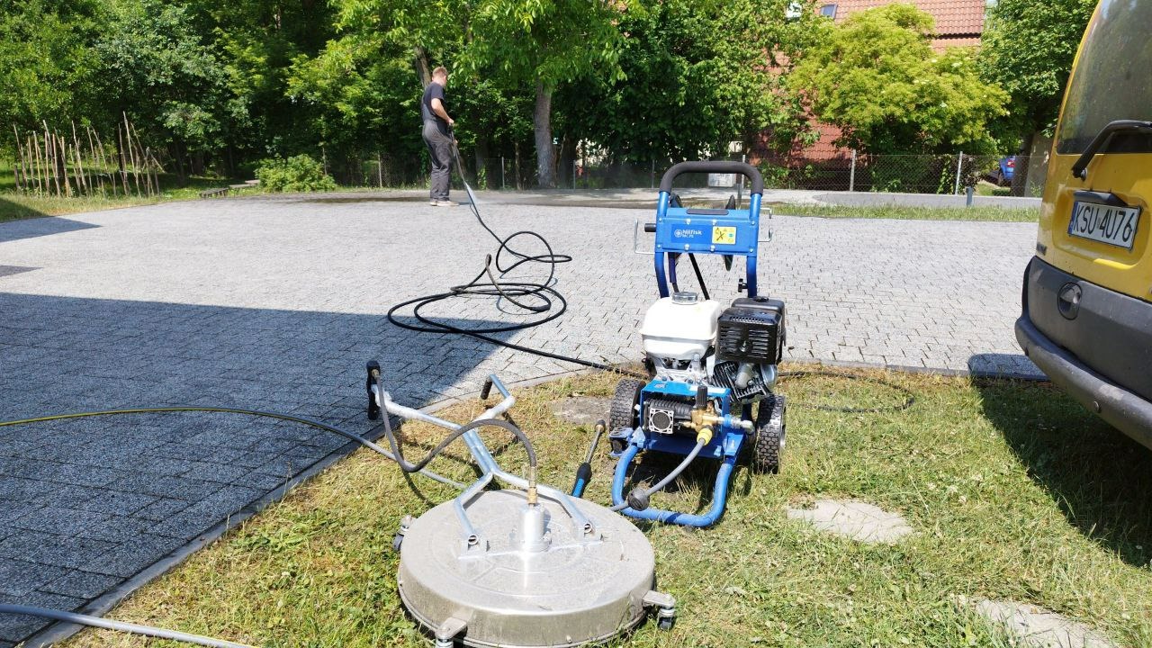 Czyszczenie kostki brukowej za pomocą myjki ciśnieniowej Nilfisk z dyszą rotacyjną i dodatkową przystawką do powierzchni płaskich. Widoczny operator w trakcie pracy oraz zaparkowany żółty samochód...
