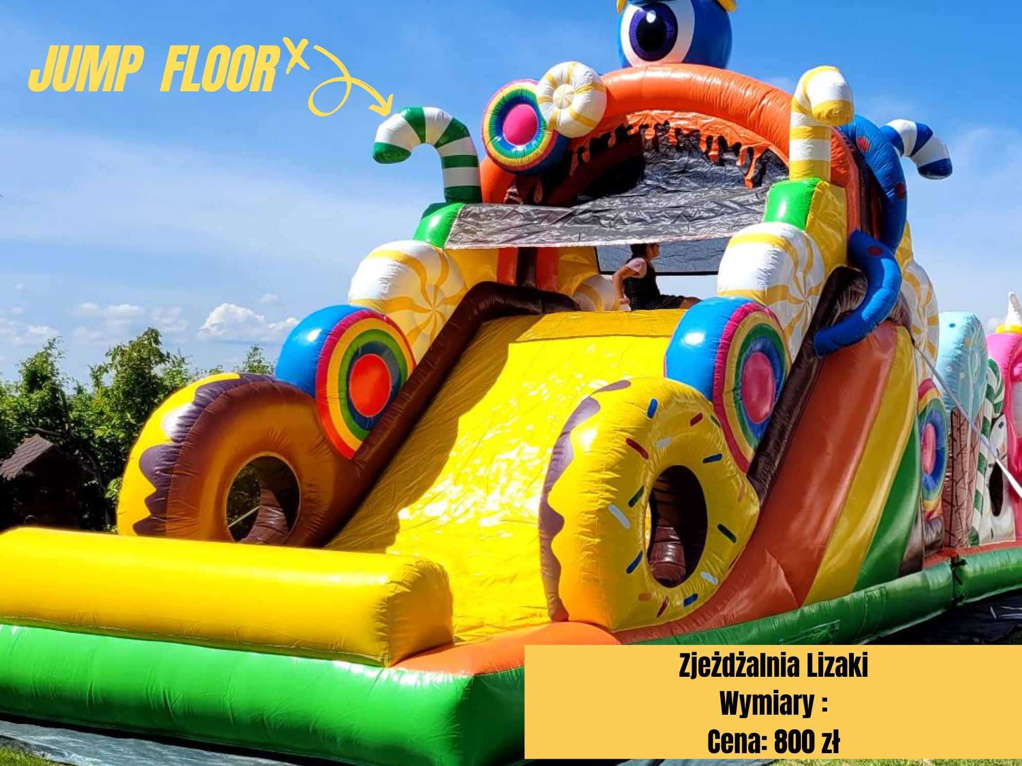 Kolorowa dmuchana zjeżdżalnia w kształcie słodyczy, z elementami imitującymi lizaki i pączki, ustawiona na trawie w słoneczny dzień. Widoczna osoba na górze zjeżdżalni.