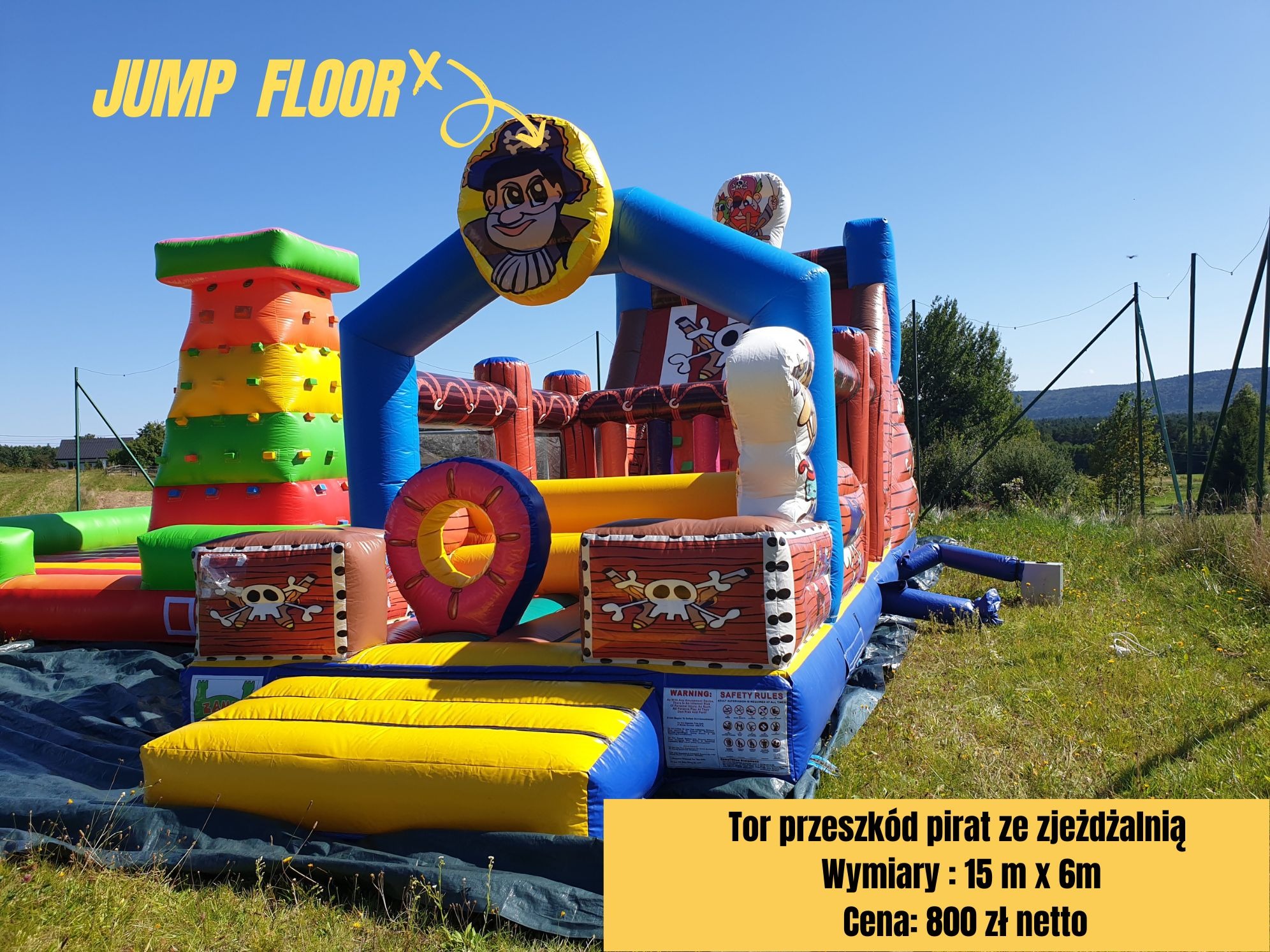 Duży, kolorowy dmuchany tor przeszkód z motywem pirackim, z elementami wspinaczkowymi i zjeżdżalnią, na tle zielonego trawnika i błękitnego nieba.