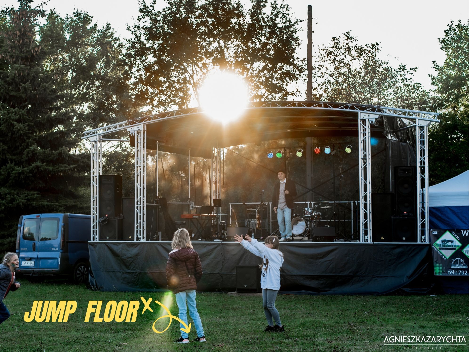 Scena plenerowa z nagłośnieniem i oświetleniem, na której stoi mężczyzna i instrumenty muzyczne, a przed nią bawi się dwoje dzieci na trawie.