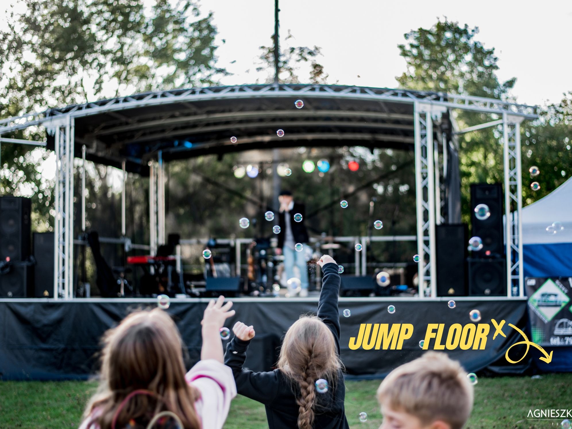 Dzieci wyciągają ręce do baniek mydlanych na tle sceny z muzykiem podczas imprezy plenerowej.