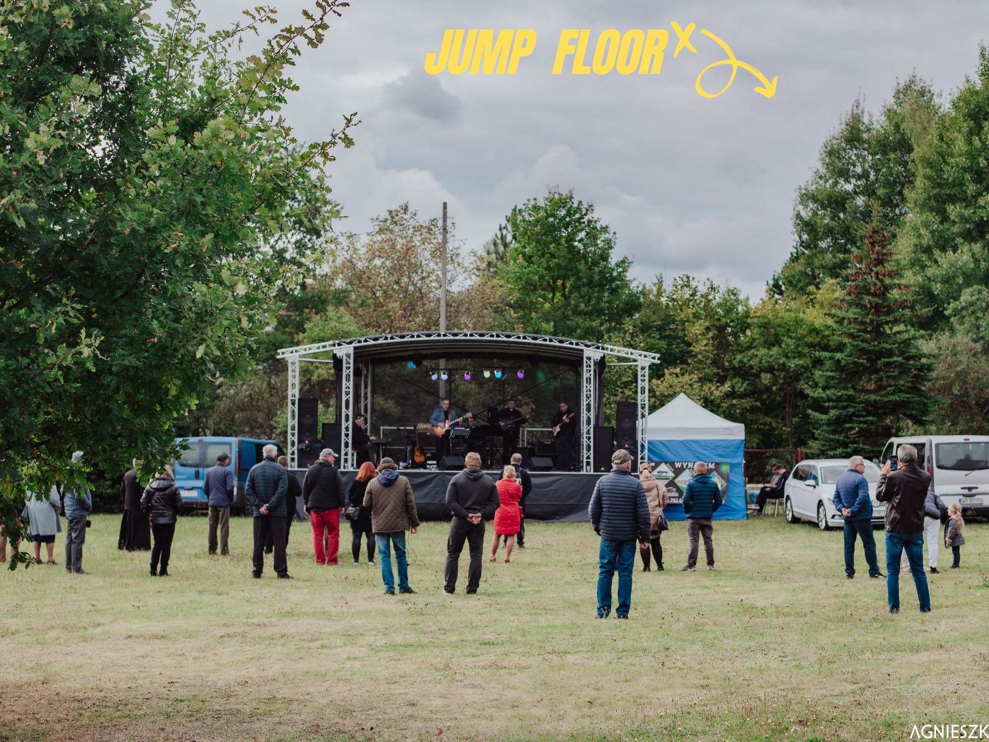 Plenerowy koncert z zespołem na scenie, publiczność na trawie, częściowo zasłonięty namiot eventowy w tle, drzewa dookoła.