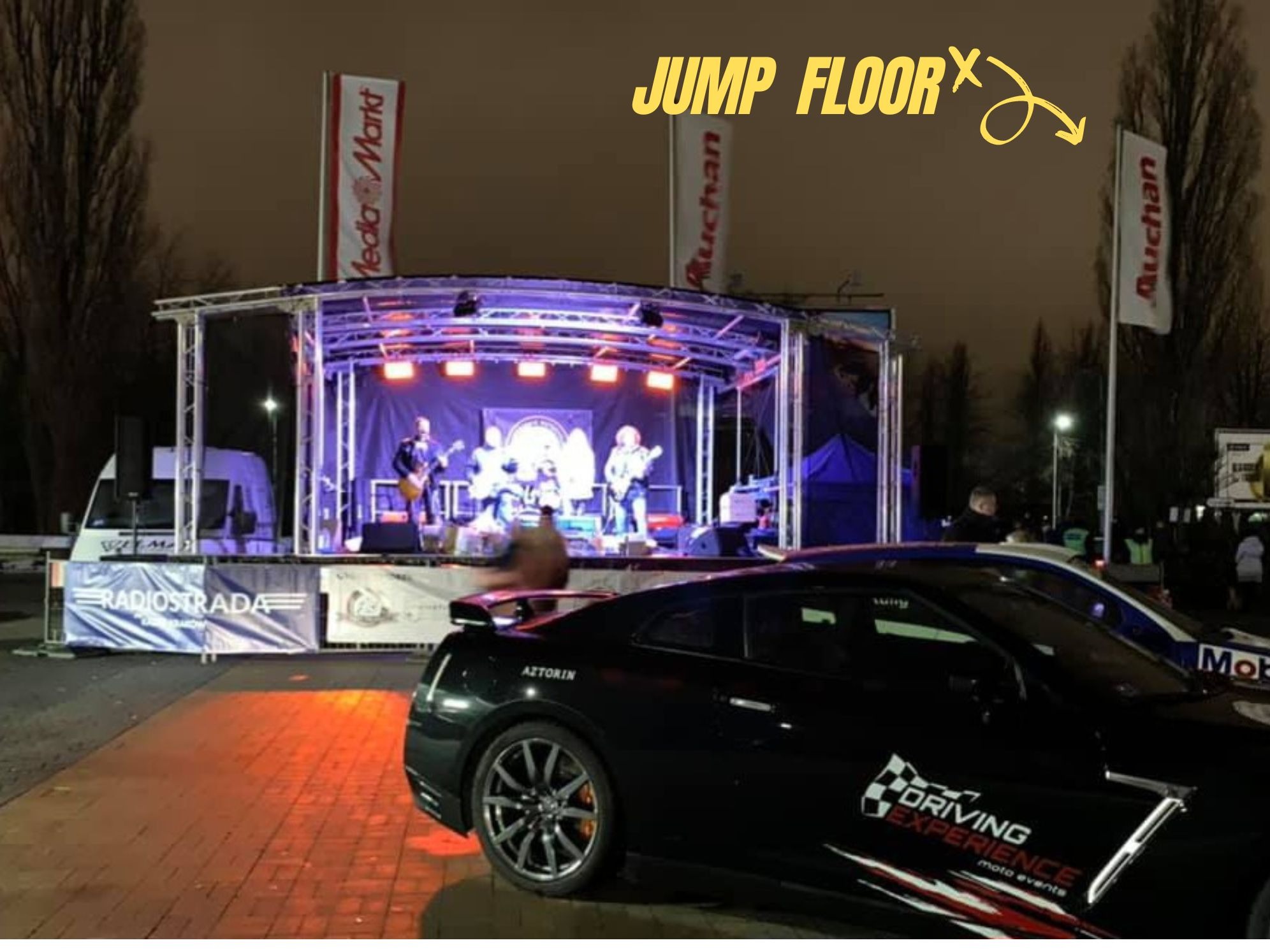 Zespół muzyczny na scenie podświetlonej na niebiesko, z flagami reklamowymi w tle, częściowo zasłonięty przez czarny samochód sportowy z naklejką 'Driving Experience'.
