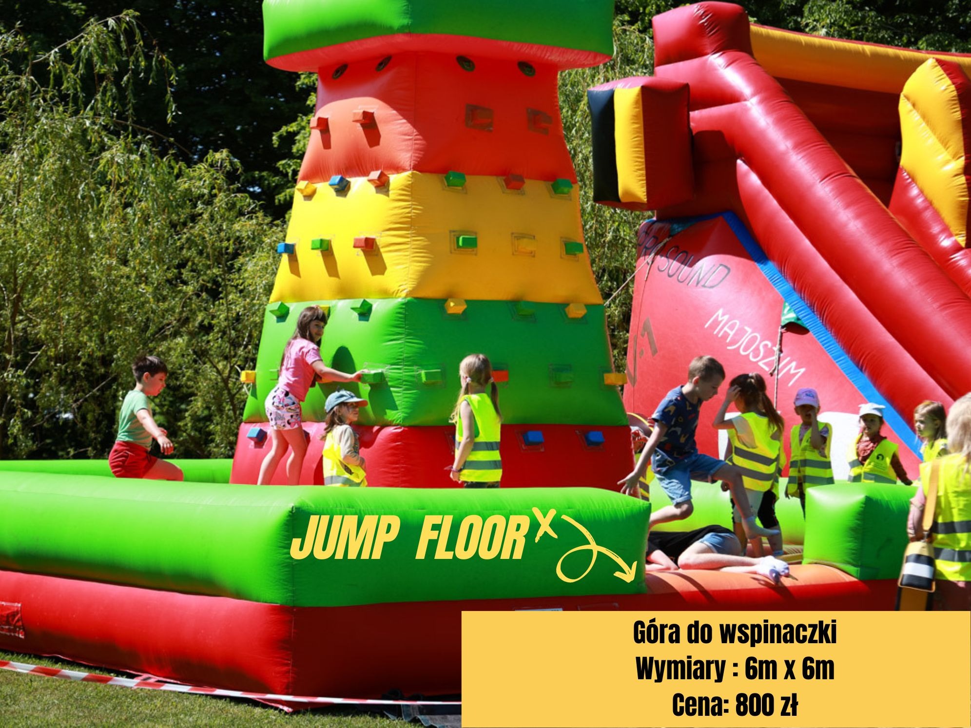 Dzieci bawią się na dmuchanym placu zabaw z kolorową ścianką wspinaczkową i zjeżdżalnią, na trawiastym terenie.