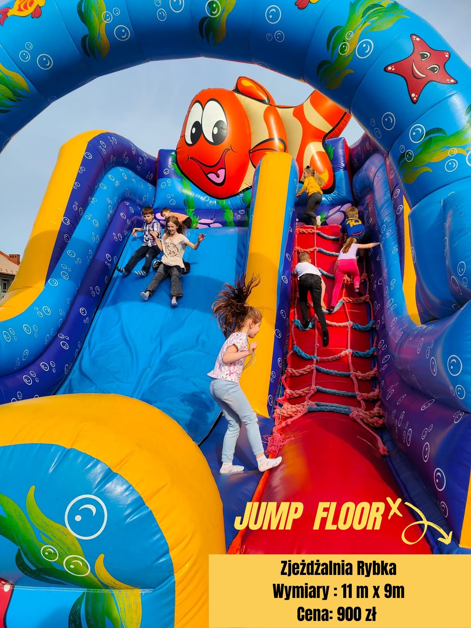 Dzieci w różnym wieku bawią się na dmuchanej zjeżdżalni w kształcie ryby z elementami w stylu morskim, wspinając się po czerwonej siatce i zjeżdżając po niebieskim ślizgu.