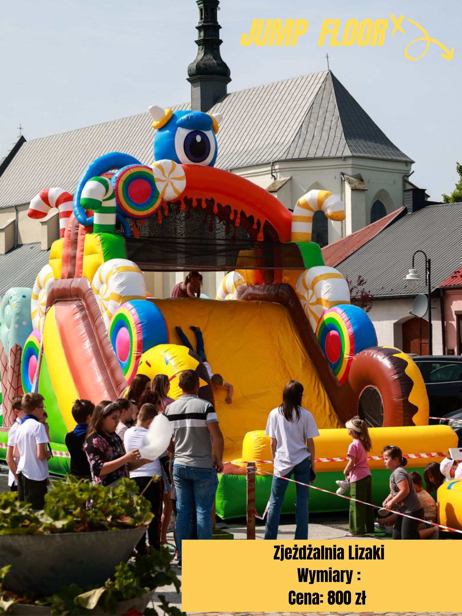 Duży, kolorowy dmuchany zamek w kształcie słodyczy z motywem lizaków i cukierków, otoczony grupą dzieci i dorosłych, na tle budynku kościoła.