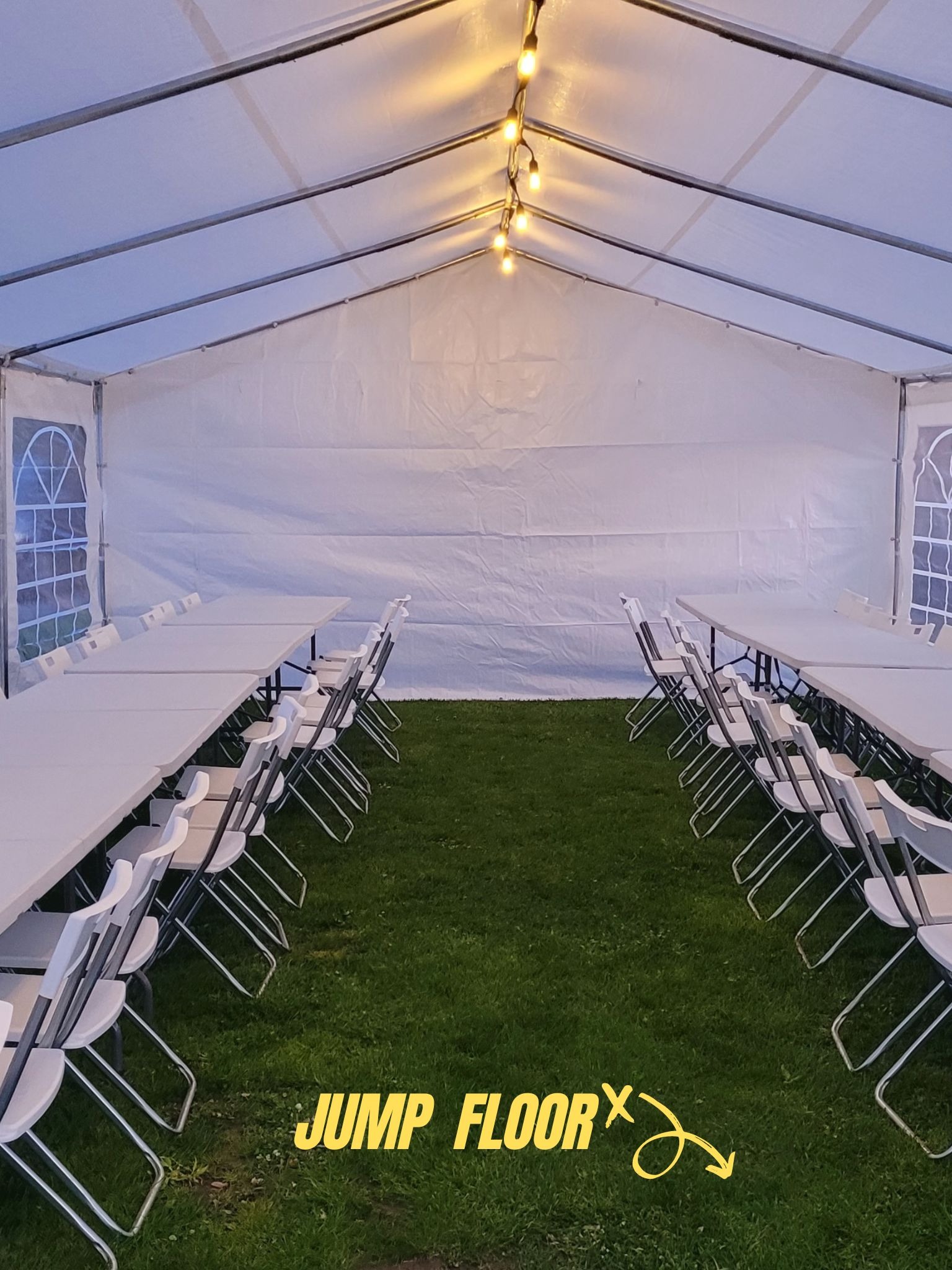 Wnętrze białego namiotu z rzędami białych stołów i krzeseł ustawionych na trawie, oświetlone sznurem żarówek