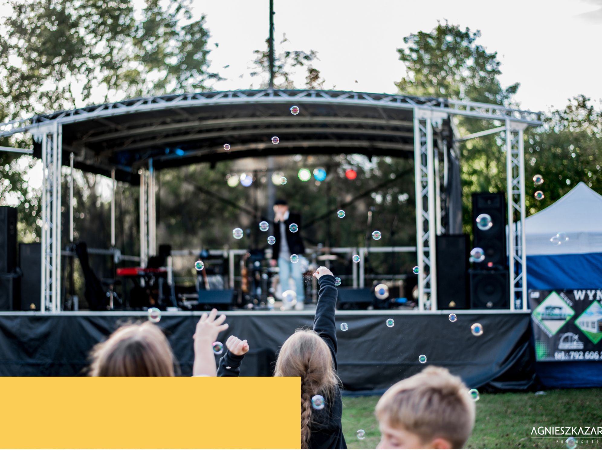 Dzieci wyciągają ręce do baniek mydlanych na tle sceny z nagłośnieniem i oświetleniem podczas imprezy plenerowej.