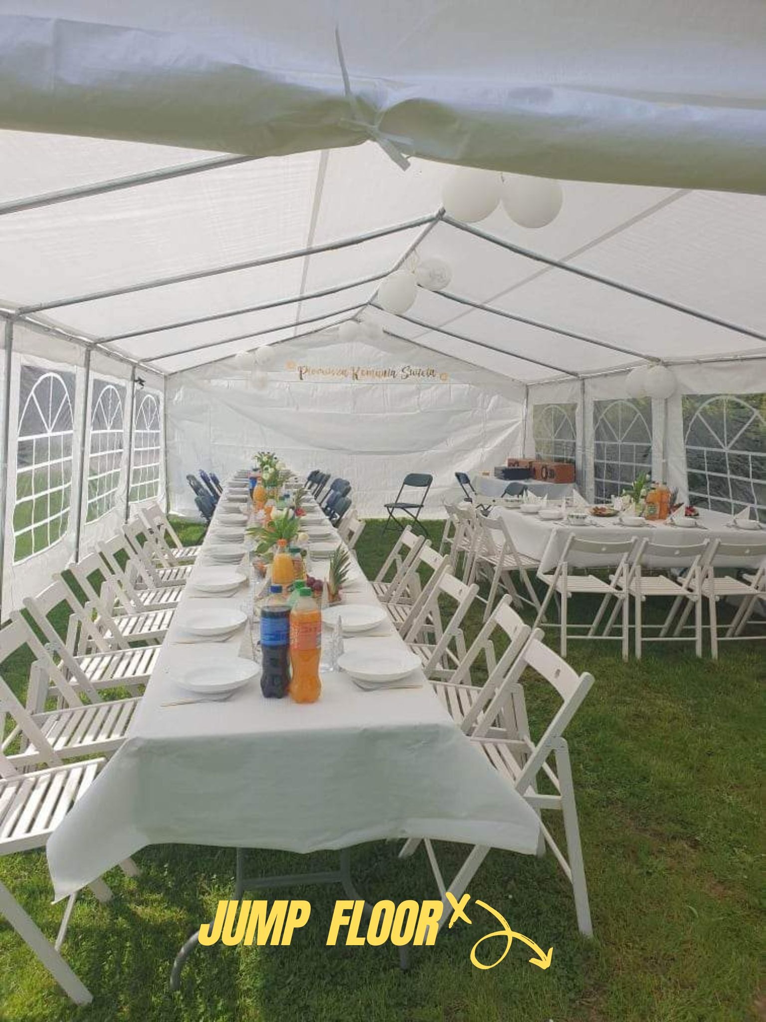 Wnętrze białego namiotu z rozstawionymi stołami i krzesłami, przygotowanego na uroczystość na trawie, z dekoracjami kwiatowymi i napojami na stołach.