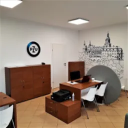 Wnętrze biura z logo PZU na ścianie, biurkiem z laptopem, drukarką i rysunkiem zamku na ścianie w tle.