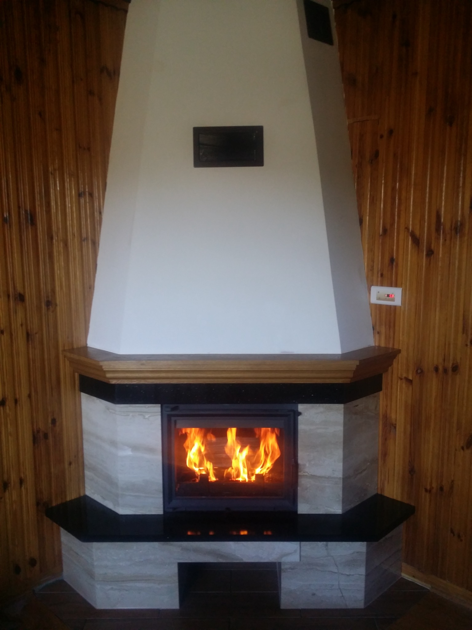 Nowoczesny kominek z obudową z jasnego marmuru, drewnianym blatem i czarnymi elementami wykończeniowymi, z palącym się ogniem wewnątrz paleniska, na tle drewnianej boazerii.