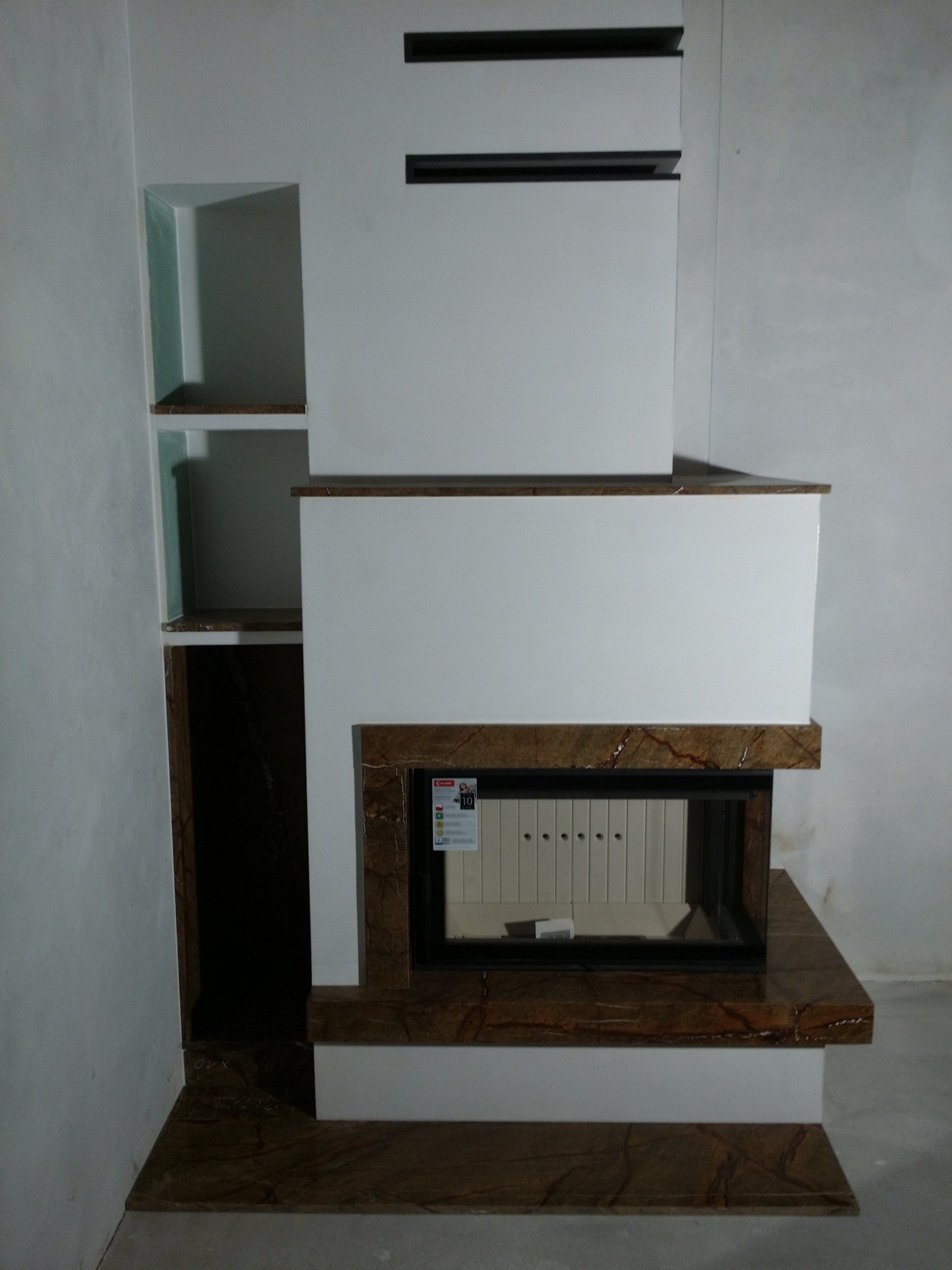 Nowoczesna obudowa z marmuru i białych płyt, z wnękami po lewej stronie, tworząca zabudowę kominkową z wkładem kominkowym.