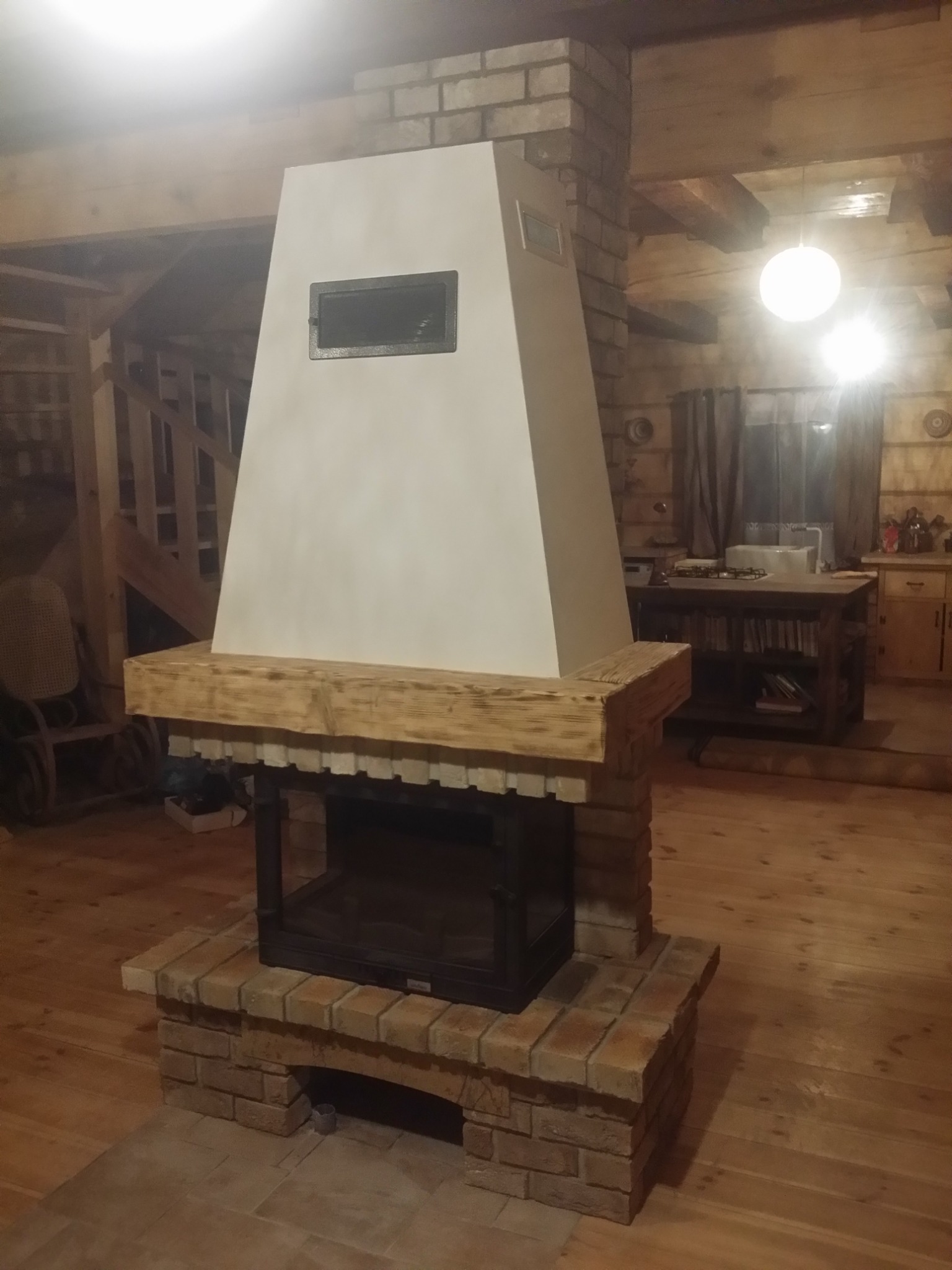 Murowany kominek z jasną, trapezową obudową i drewnianą belką w drewnianym wnętrzu domu.