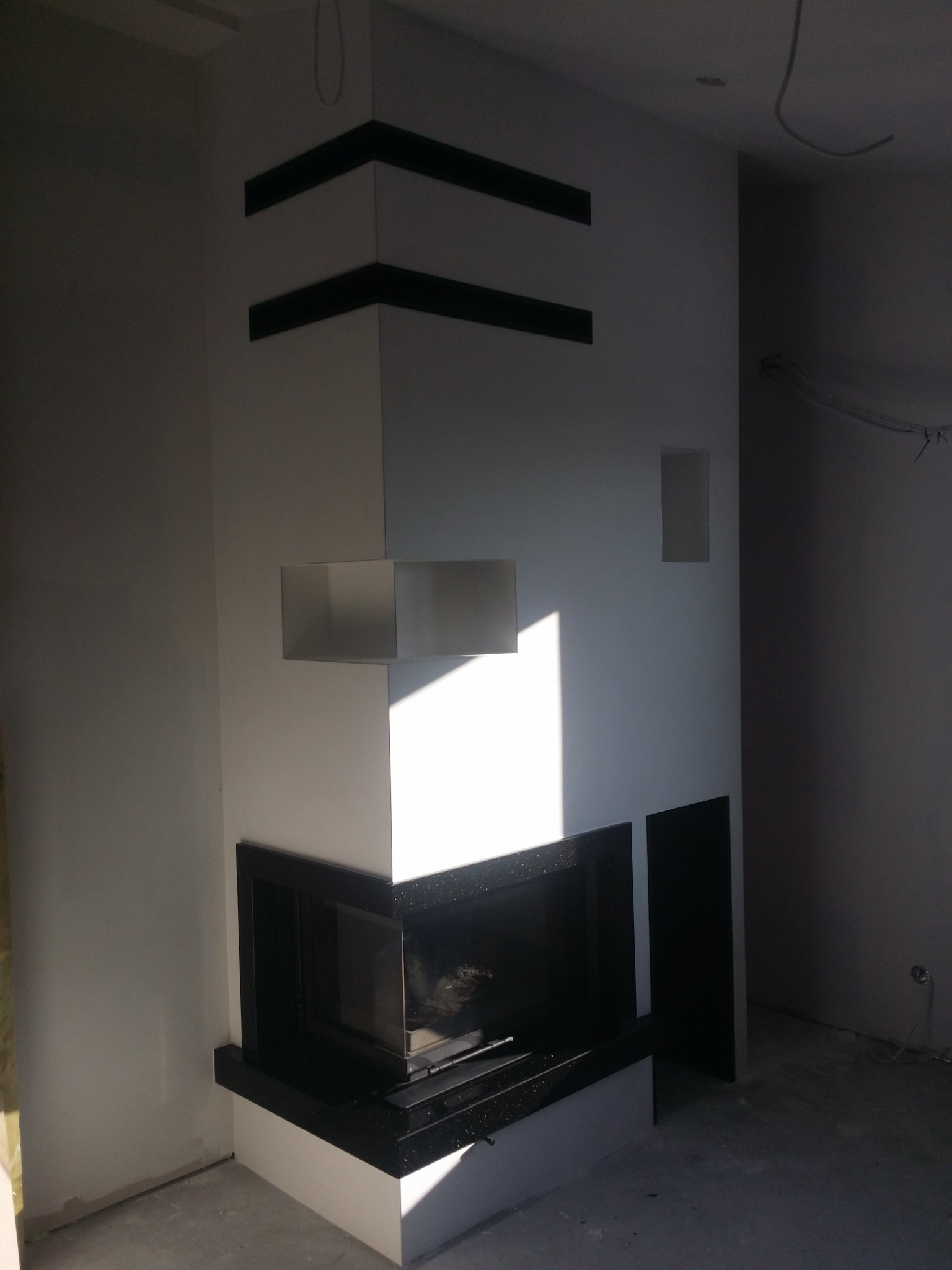 Nowoczesny wkład kominkowy z czarną obudową i elementami dekoracyjnymi na białej ścianie w surowym wnętrzu.