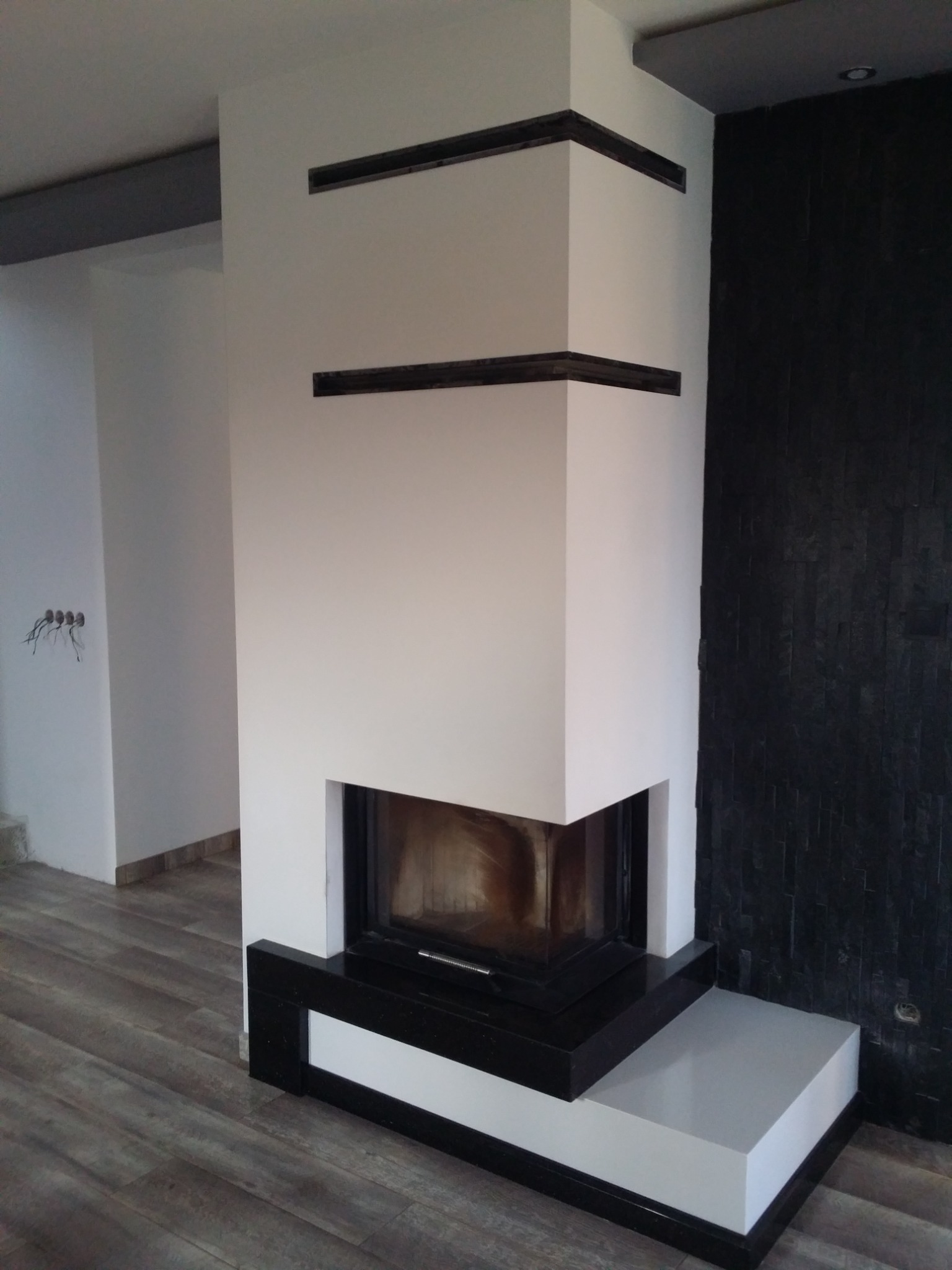 Nowoczesny, biało-czarny wkład kominkowy z dekoracyjnymi czarnymi listwami na ścianie, ustawiony na dwupoziomowej podstawie, z ciemną okładziną ściany w tle i drewnopodobną podłogą.