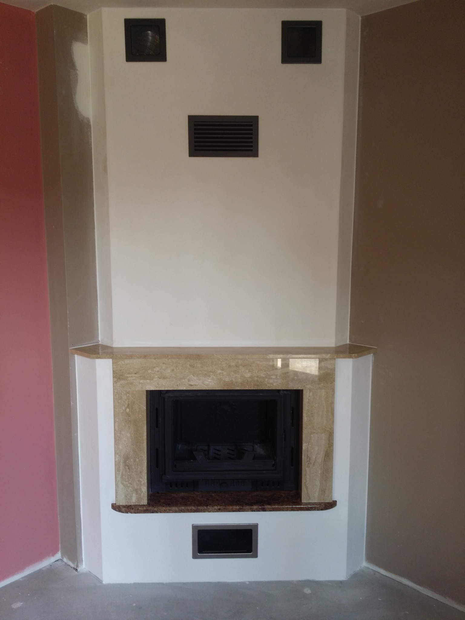 Obudowa wkładu kominkowego wykończona trawertynem, z trzema kratkami wentylacyjnymi w górnej części i jedną u dołu, na tle ścian w odcieniach różu i beżu.