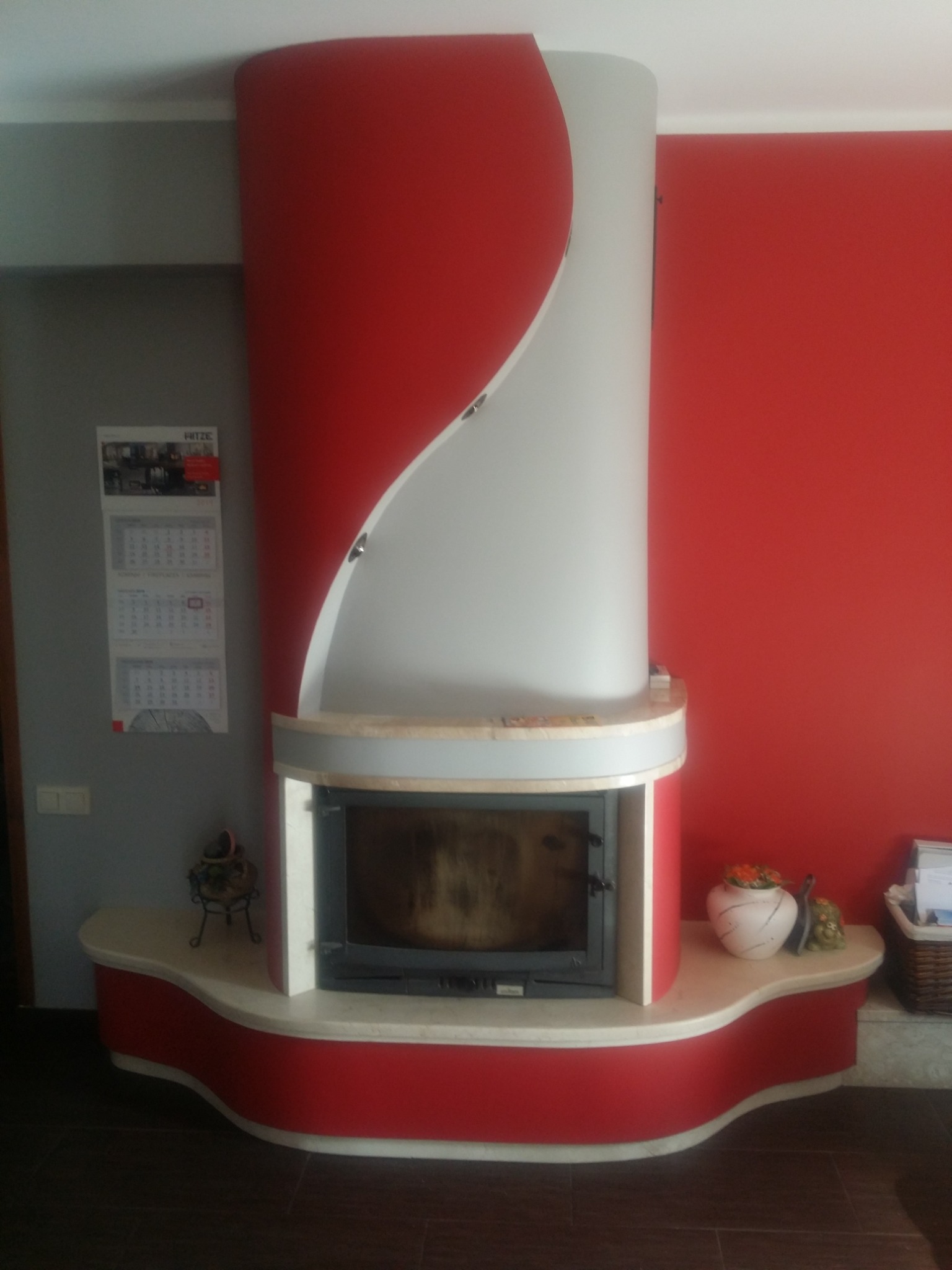 Nowoczesny, cylindryczny model obudowy z elementami w kolorze czerwonym i szarym, z zamkniętym paleniskiem, na tle czerwonej ściany i kalendarza.