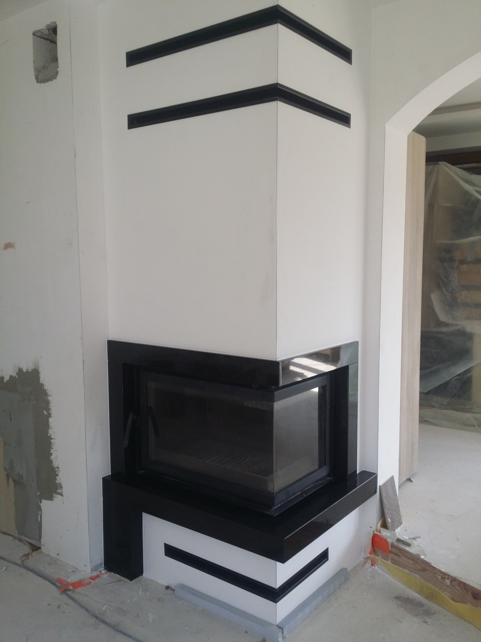 Nowoczesny narożny wkład kominkowy z czarną obudową, w trakcie montażu, widoczne niedokończone prace wykończeniowe na ścianie.